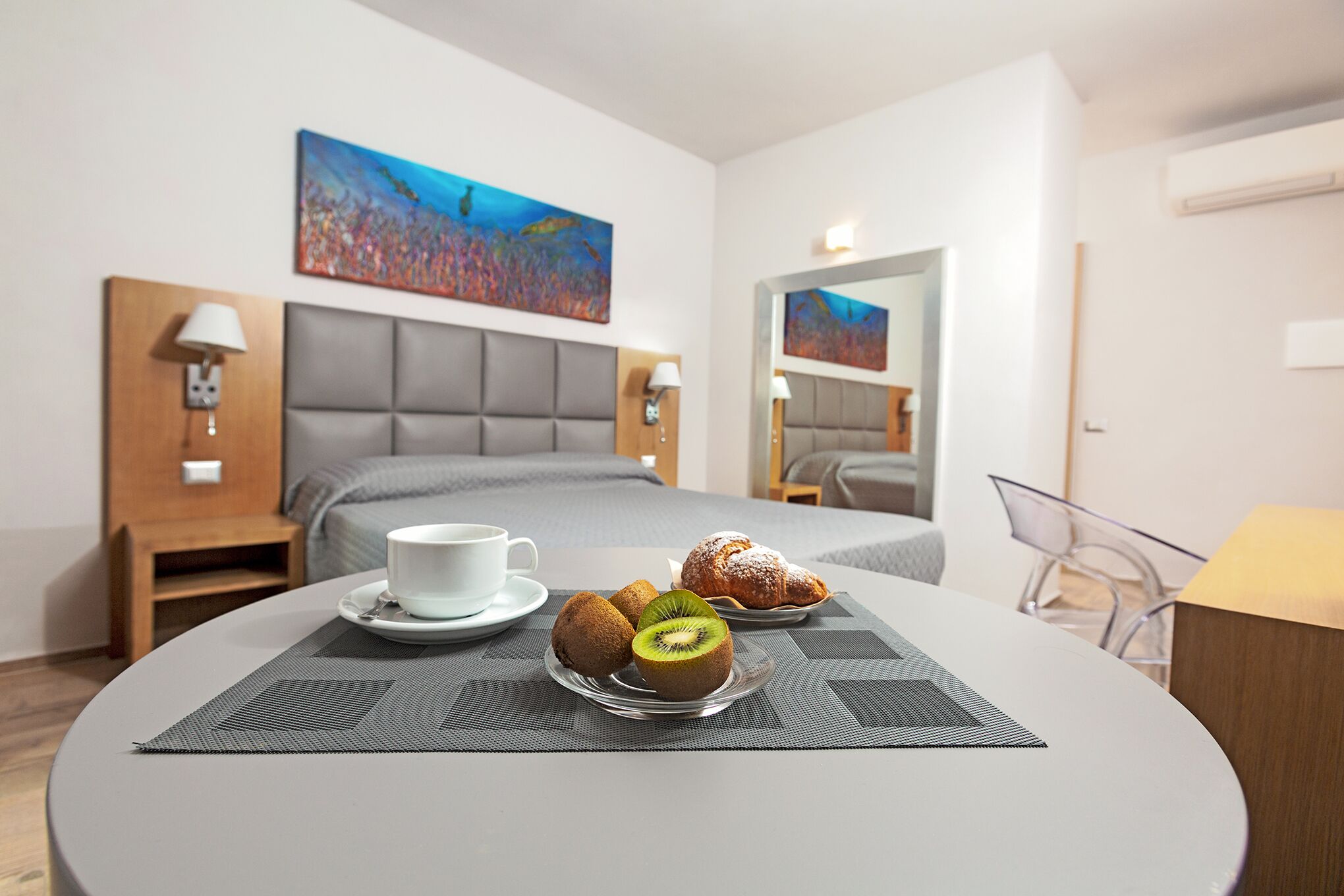 Quarto de hotel com uma cama, uma mesa de jantar com pastelaria e um kiwi, uma chávena branca e um quadro na parede.