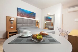 A hotel room with a bed, a dining table with pastries and a kiwi, a white cup, and a painting on the wall. - Capotesta Luxury Rooms (Santa Teresa di Gallura)