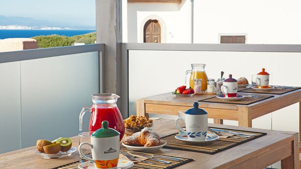 Ett frukostbord med croissanter, ett glas apelsinjuice och en kanna med röd juice på en balkong med utsikt över kusten.