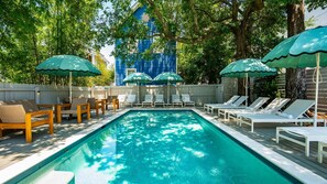 A swimming pool with lounge chairs and umbrellas, surrounded by trees and a blue building in the background. - The Iris, Signature Suite (Charleston)
