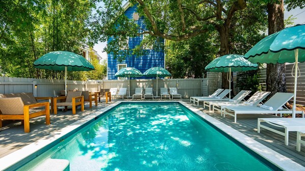 A swimming pool with lounge chairs and umbrellas, surrounded by trees and a blue building in the background.