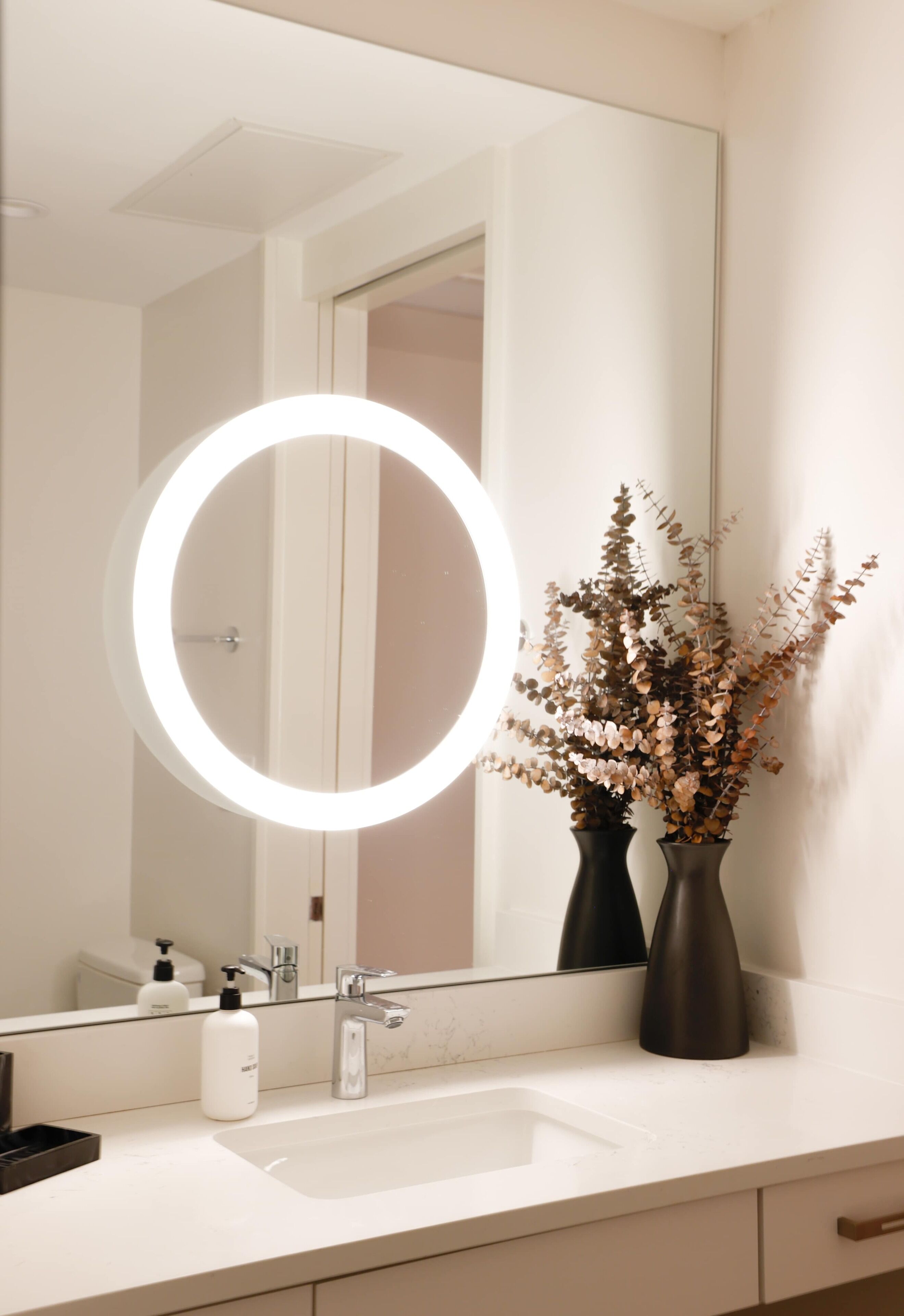 A bathroom with a round mirror, a white sink, and two dark vases with dried flowers.
