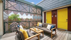Een houten terras met rieten meubilair, een tafel met een schaal eten, en een gele deur.