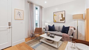 Een moderne woonkamer met een grijze bank, een witte salontafel en een bruine fauteuil. Er hangt een lijst met een afbeelding aan de muur, een lamp en een deur.