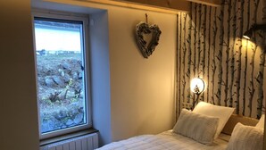 Un dormitorio con un ventanal grande, una cama con ropa de cama blanca, un adorno con forma de corazón en la pared y una lámpara de pared.