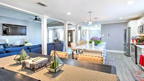 Uma cozinha moderna com eletrodomésticos de aço inoxidável, uma sala de jantar com mesa de madeira e uma sala de estar com sofá azul e plantas decorativas.