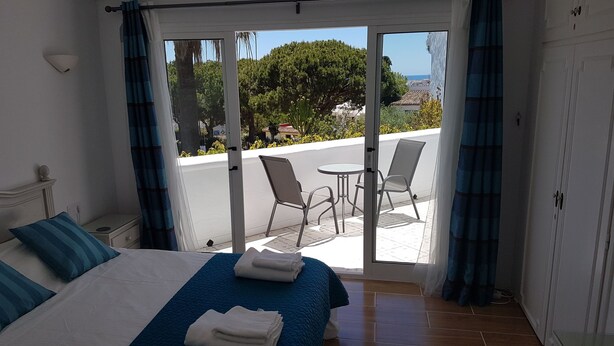 A hotel room with a bed, a small table, two chairs, and a view of the street and greenery outside. - Holiday Home "Casa Los Pinos" with Sea View, Shared Pool, Balcony, A/C & Wi-Fi (Sitio de Calahonda)