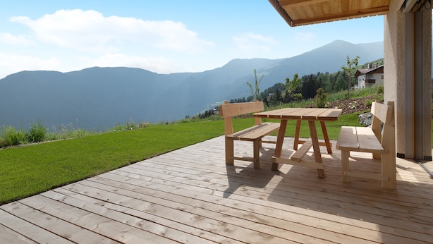 Um deck de madeira com uma mesa e bancos, com vista para uma área gramada e montanhas.