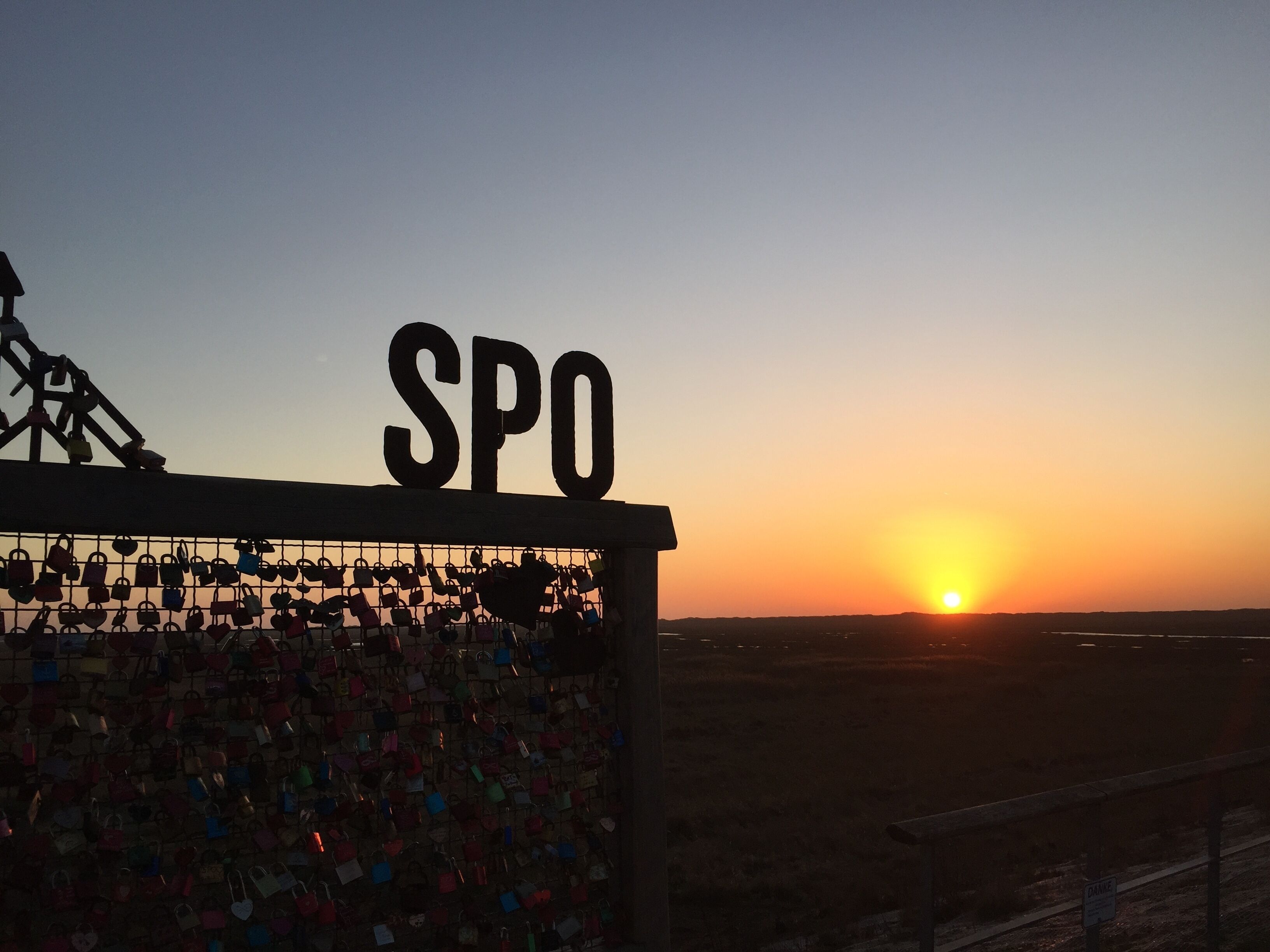 Blick auf einen Sonnenuntergang mit einem Schild mit der Aufschrift 'SPO' und einer Mauer voller Vorhängeschlösser.