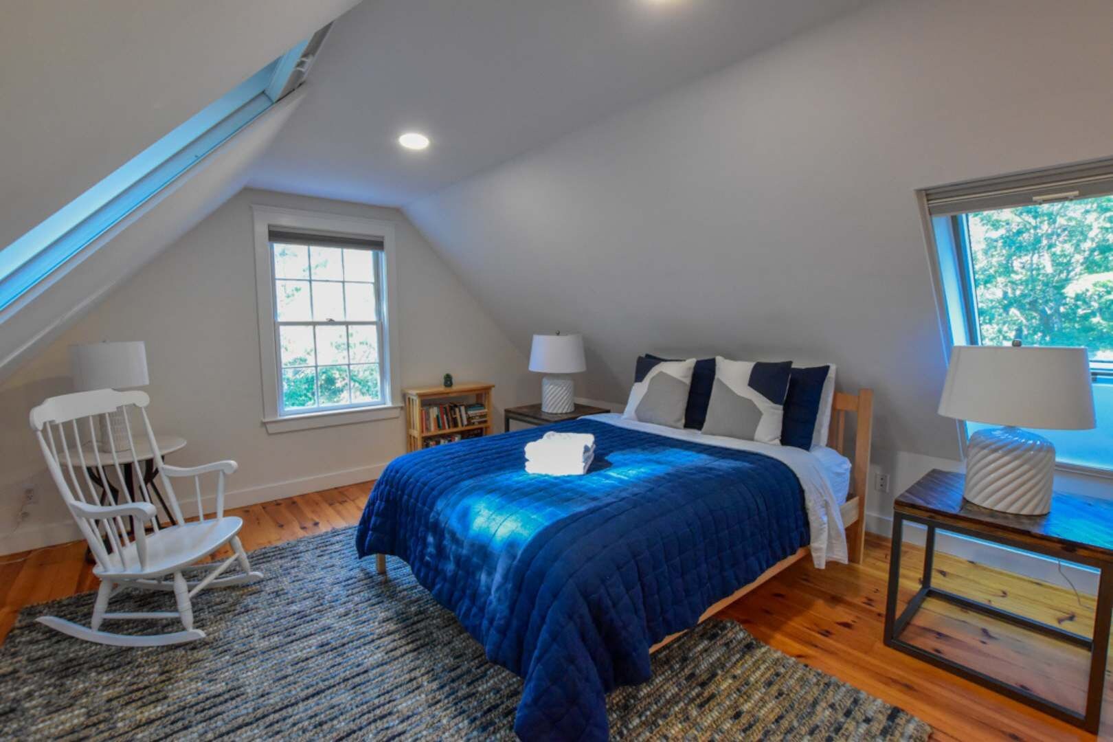 Une chambre avec un plafond en pente, un lit recouvert d’une couverture bleue, un fauteuil à bascule, une petite table de chevet avec une lampe et une fenêtre donnant sur les arbres.