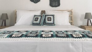 Una cama con colcha blanca y dos almohadones de crochet, cabecera de madera y dos tapices de macramé en la pared.