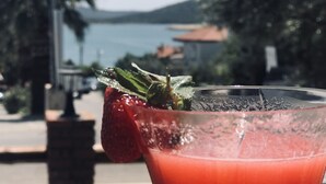 Una mano sostiene un vaso con una bebida roja y una decoración de fresa, con vistas panorámicas al agua y follaje verde de fondo.