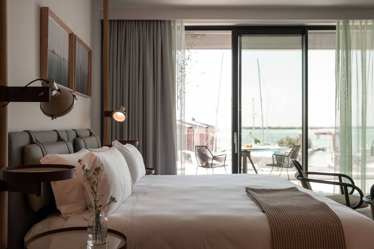 A hotel room with a large bed, a bedside table with a lamp, a vase with flowers, and a view of the sea through large windows.