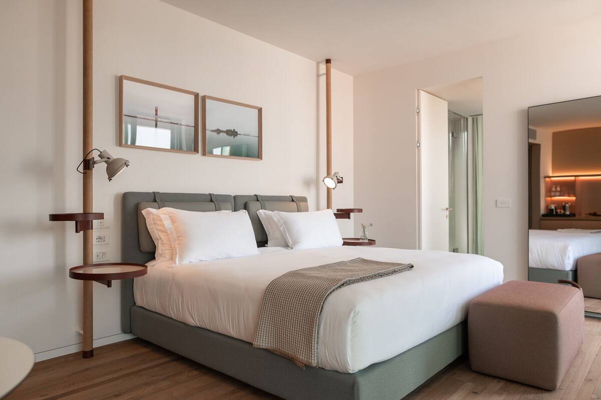 A modern hotel room with a large bed, bedside tables, a nightstand, a bedside lamp, a mirror, and a small table.