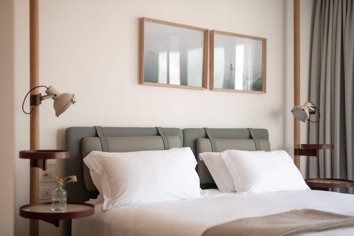A modern hotel room with a large bed, two bedside tables, a small vase with flowers, and two wall-mounted lamps.