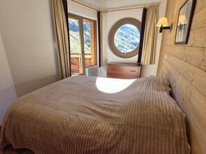 A bedroom with a large bed, a wooden headboard, a nightstand, a dresser, a window with a view of mountains, and a round window above the dresser.