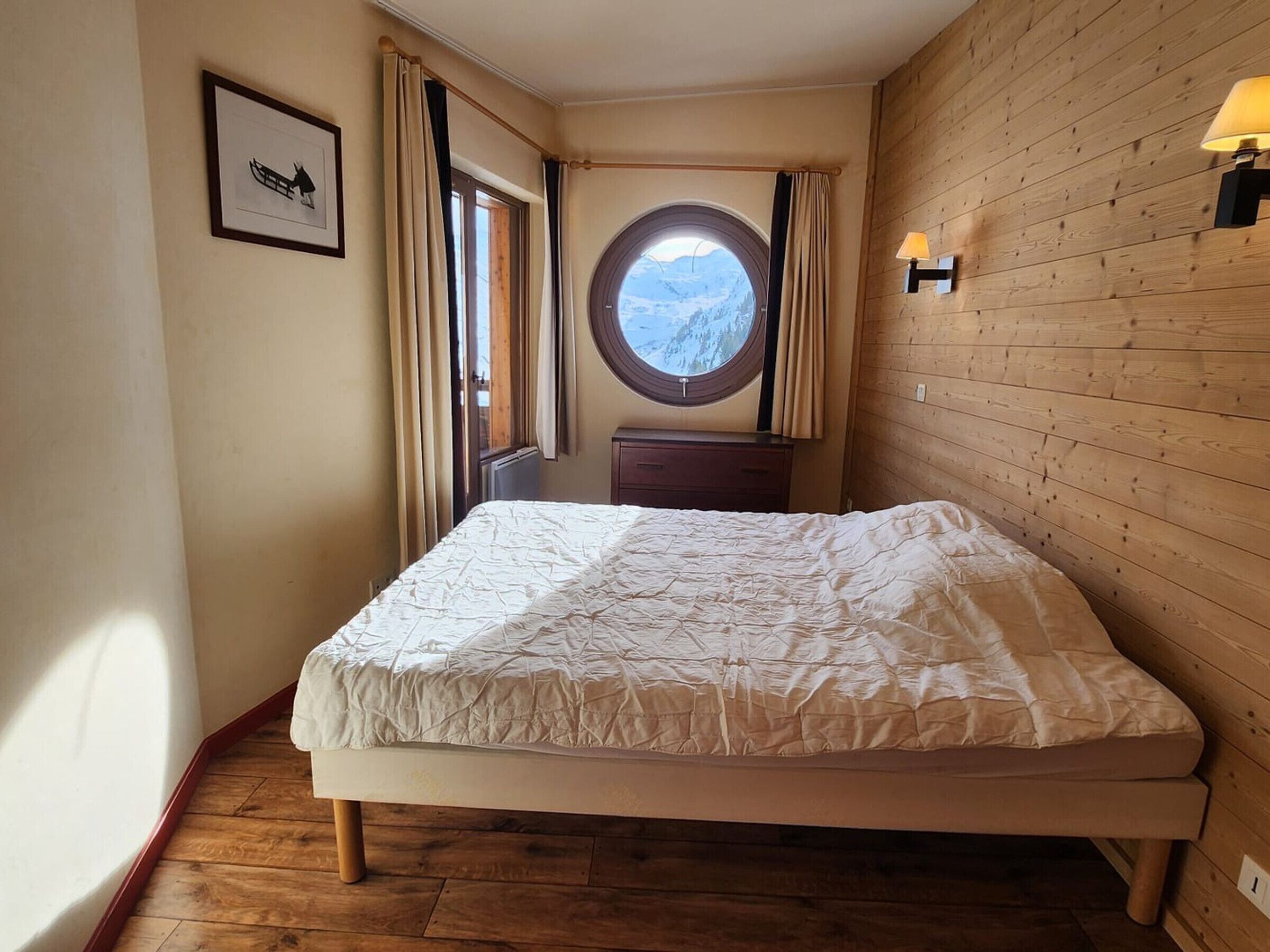 A bedroom with a wooden floor, a bed with a white duvet, a round window with a mountain view, a wooden dresser, and a wall-mounted lamp.