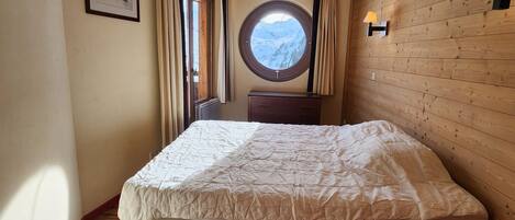 A bedroom with a wooden floor, a bed with a white duvet, a round window with a mountain view, a wooden dresser, and a wall-mounted lamp.
