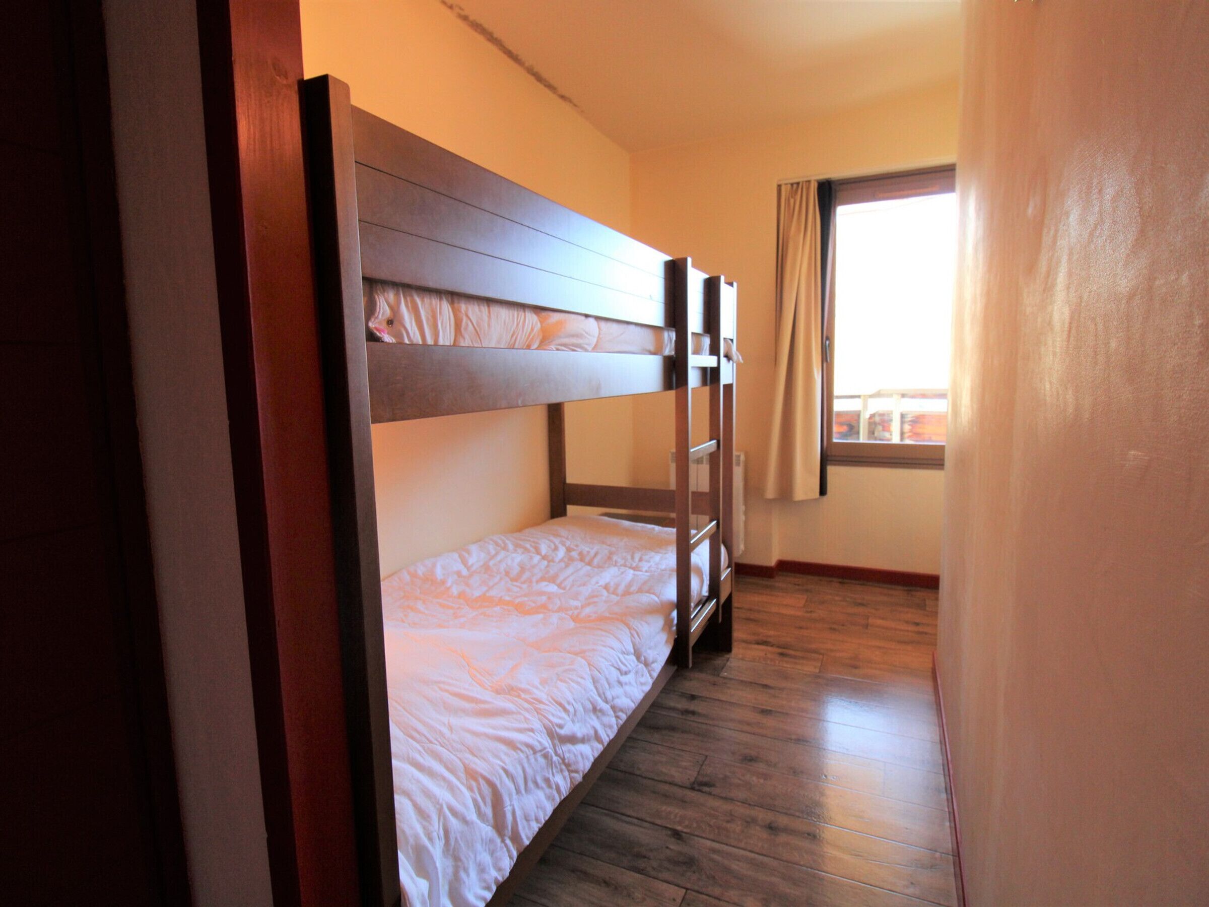 A bunk bed room with wooden headboards, a window with curtains, and a wooden floor.