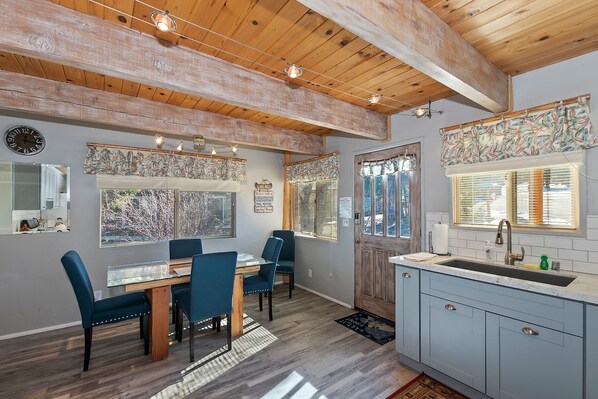 A dining area with a wooden table and chairs, a kitchen with a sink and cabinets, and a window with blinds.