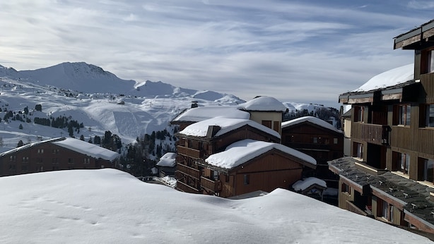 Ein verschneites Bergdorf mit Holzhäusern und einer Skipiste im Hintergrund.
