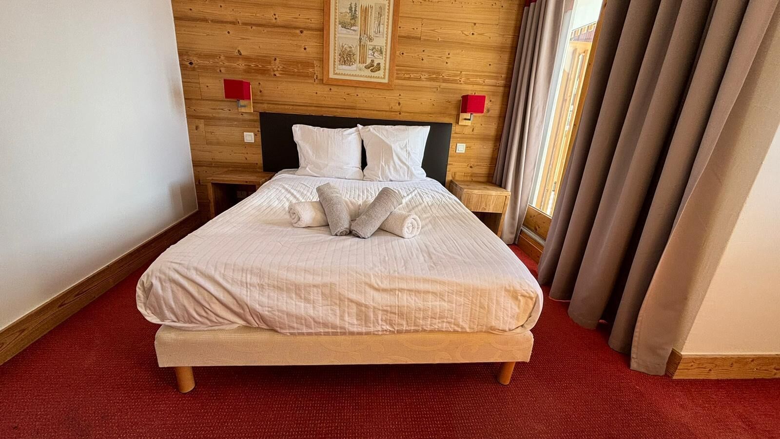 Un lit bien fait, avec du linge de lit blanc et des oreillers, une tête de lit en bois et un sol recouvert d’une moquette rouge.