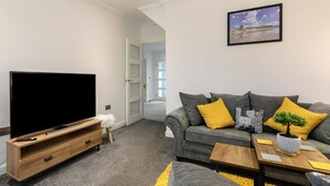 A modern living room with a flat-screen TV on a wooden stand, a grey sofa with yellow cushions, a small table with a plant, and a framed picture on the wall. - Ocean Waves - Ground Floor Apartment Pet Friendly Enclosed Garden (Saundersfoot)