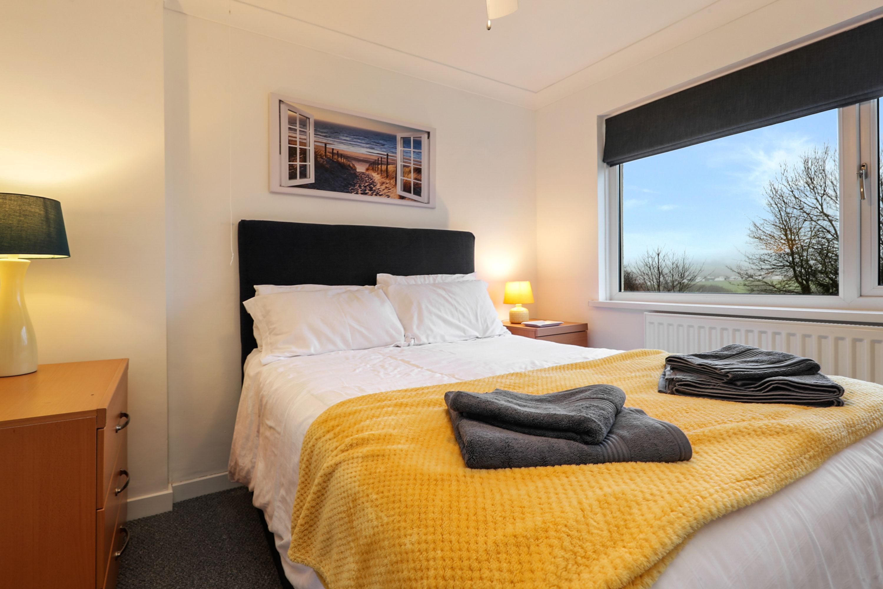 A neatly made bed with a yellow blanket, a black headboard, and white pillows. There is a framed picture above the bed, a lamp on the nightstand, and a window with a view of the outdoors.