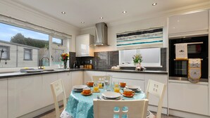 A modern kitchen with white cabinets, a stainless steel range hood, and a dining area set with a blue tablecloth and white chairs. - Plas Gwyn - Walk to Beach Parking Enclosed Garden (Saundersfoot)