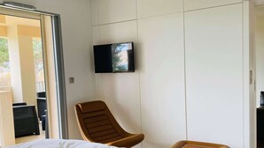 A modern bedroom with a large bed, a brown chair, a small stool, and a television mounted on the wall. - 3-room apartment, 2 beds, beach, parking, AC, WiFi (Calvi)