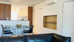 A modern living room with a black leather sofa, a dining area with a table and chairs, and a kitchen area with wooden cabinets and an air conditioning unit. - 3-room apartment, 2 beds, beach, parking, AC, WiFi (Calvi)