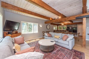 A spacious living room with a large gray sofa, a round coffee table, and a television. There is a wooden ceiling and a view of the kitchen in the background.