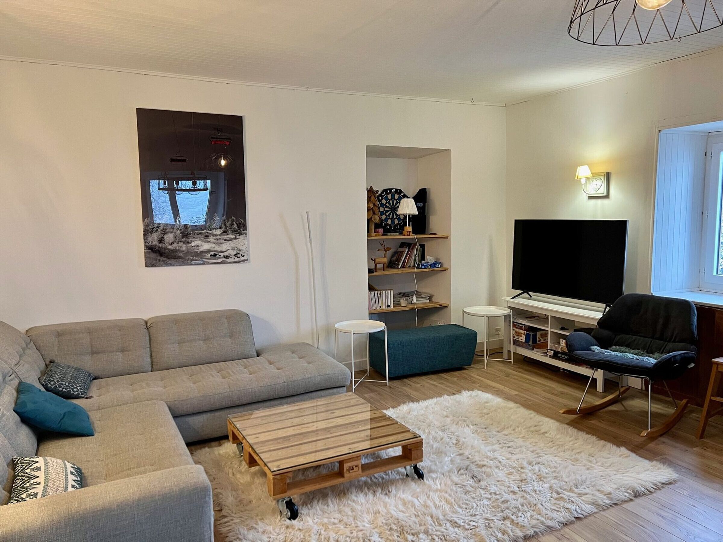 A modern living room with a grey sectional sofa, a wooden coffee table, a white furry rug, a bookshelf, a TV, and a rocking chair.
