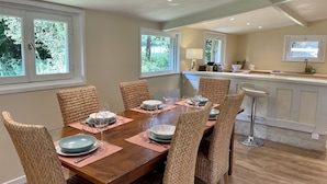 Een eetgedeelte met een houten tafel voor zes personen, omringd door rieten stoelen, en een keuken met een bargedeelte en een raam.