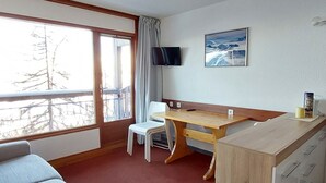 A hotel room with a wooden desk, a television mounted on the wall, a window with curtains, and a bed with a grey headboard. - 2-room apartment for 4 people in Arc 1800, balcony and equipped kitchen (Les Arcs 1800)