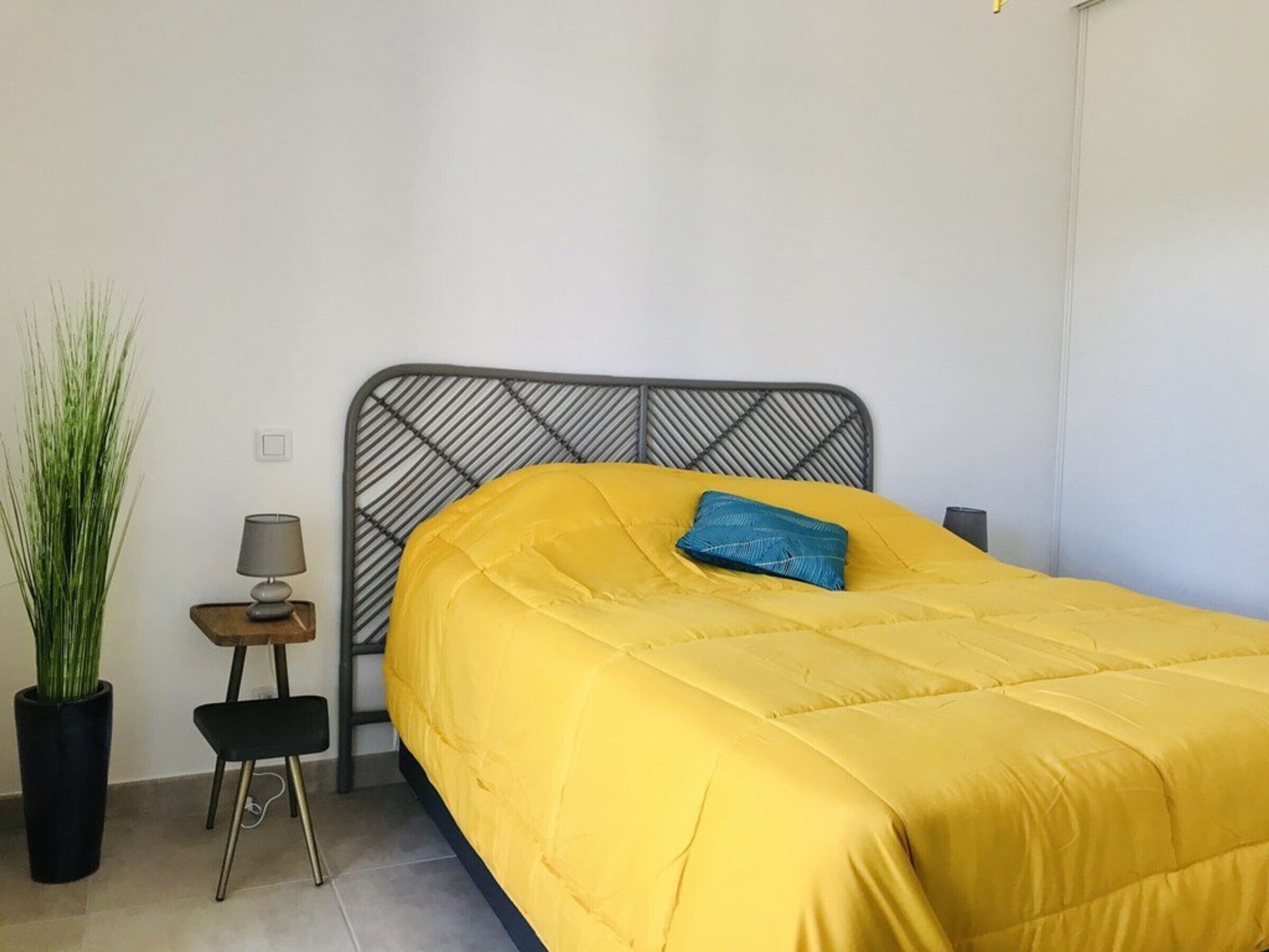 Una cama con cabecera de metal, cubrecama amarillo y almohada azul. Se observa una mesita de noche con una lámpara y una planta en maceta.