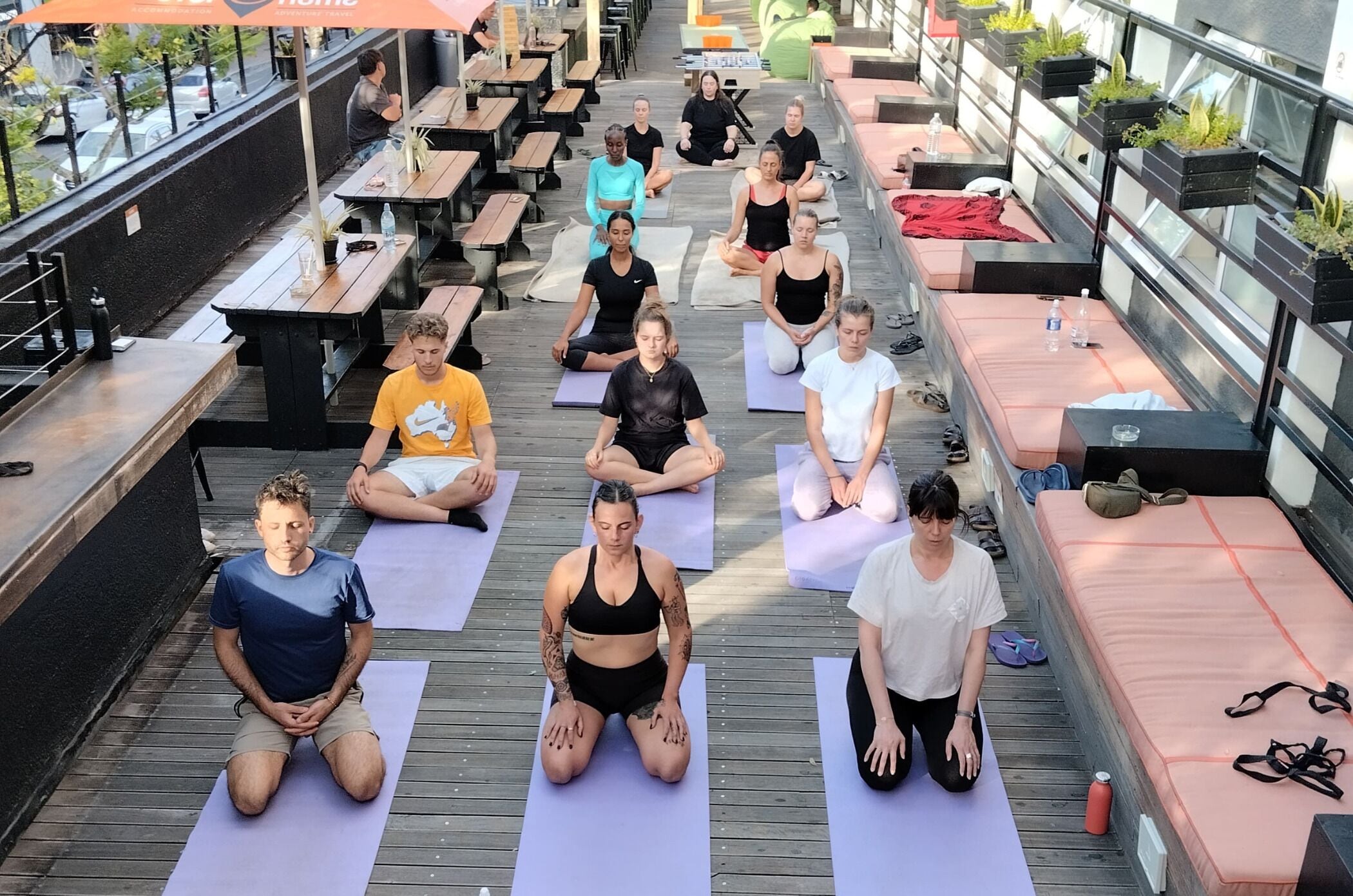 A group of people seated on yoga mats in an outdoor setting with wooden flooring, arranged in rows.