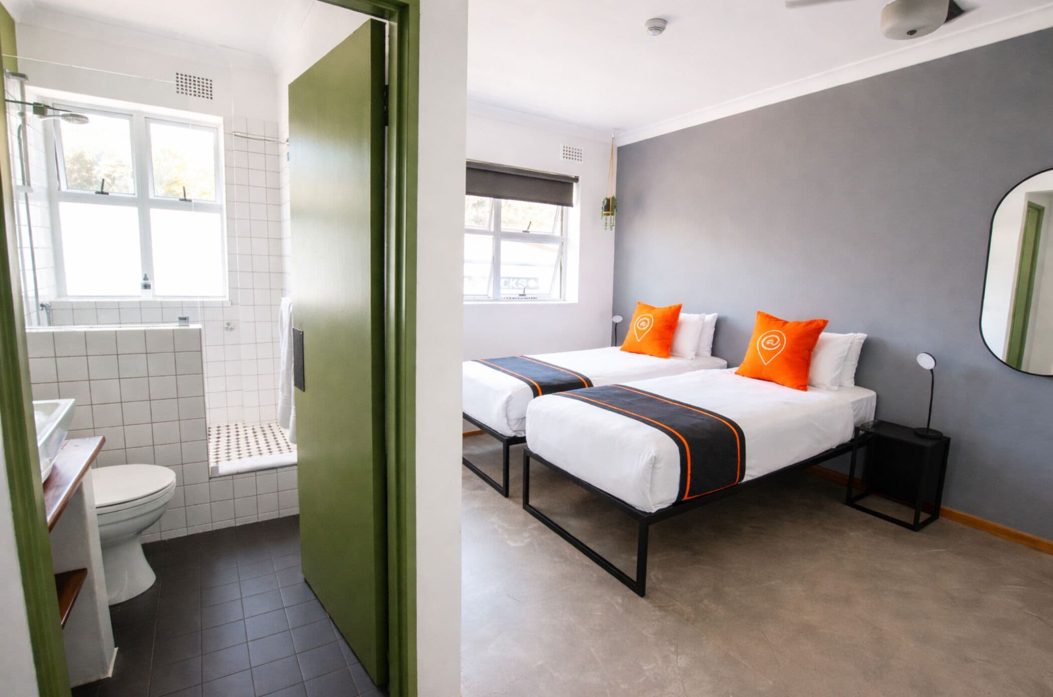A hotel room with a bathroom, two beds with orange and white bedding, a small bedside table, and a large mirror.