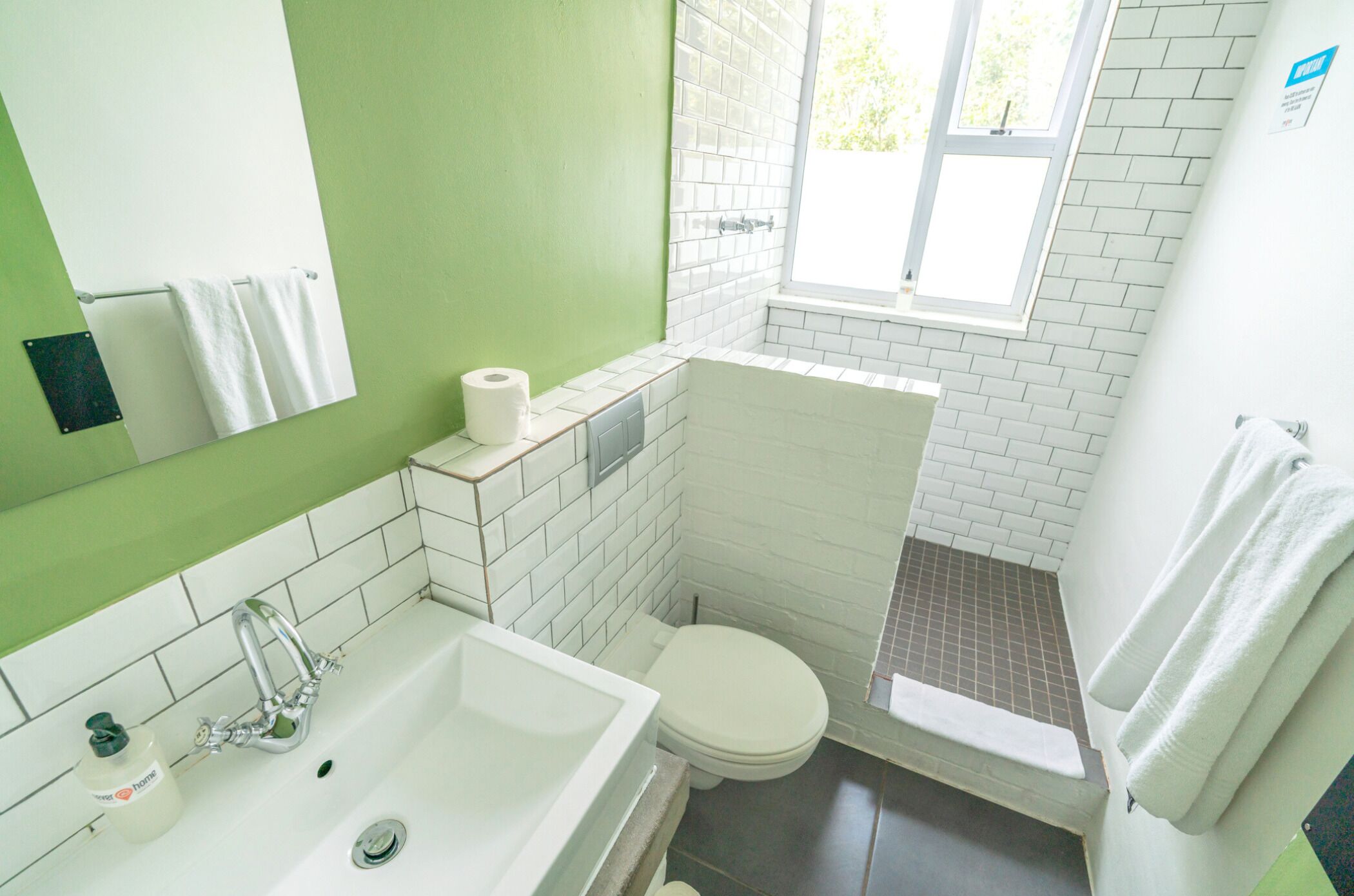 Ein Badezimmer mit weißem Waschbecken, Toilette und Duschbereich. Ein Fenster sorgt für natürliches Licht, und ein Handtuchhalter mit Handtüchern ist vorhanden.