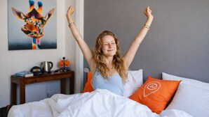 Iemand zit op een bed met witte lakens en oranje kussens, in een kamer met een grijze muur en een schilderij van een giraffe.