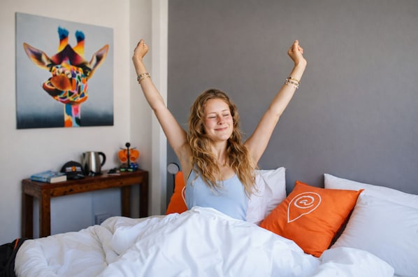 A person sitting on a bed with white bedding and orange pillows, in a room with a grey wall and a painting of a giraffe.