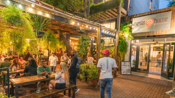 Een terras met houten tafels en banken, omgeven door potplanten en hangende lampen.