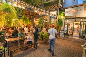An outdoor seating area with wooden tables and benches, surrounded by potted plants and hanging lights.