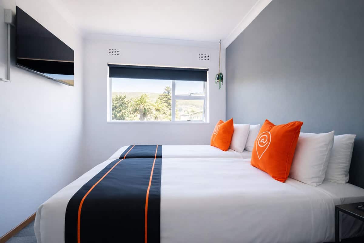 Ein Hotelzimmer mit einem großen Bett, weißer Bettwäsche, orangen Kissen, einem flachbildfernseher an der Wand und einem Fenster mit Blick auf Palmen.