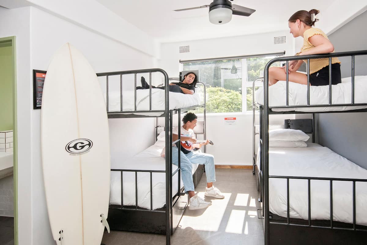 Ein Schlafsaalzimmer mit Stockbetten, einem an der Wand stehenden Surfbrett und einem Fenster mit Blick auf Grünflächen.