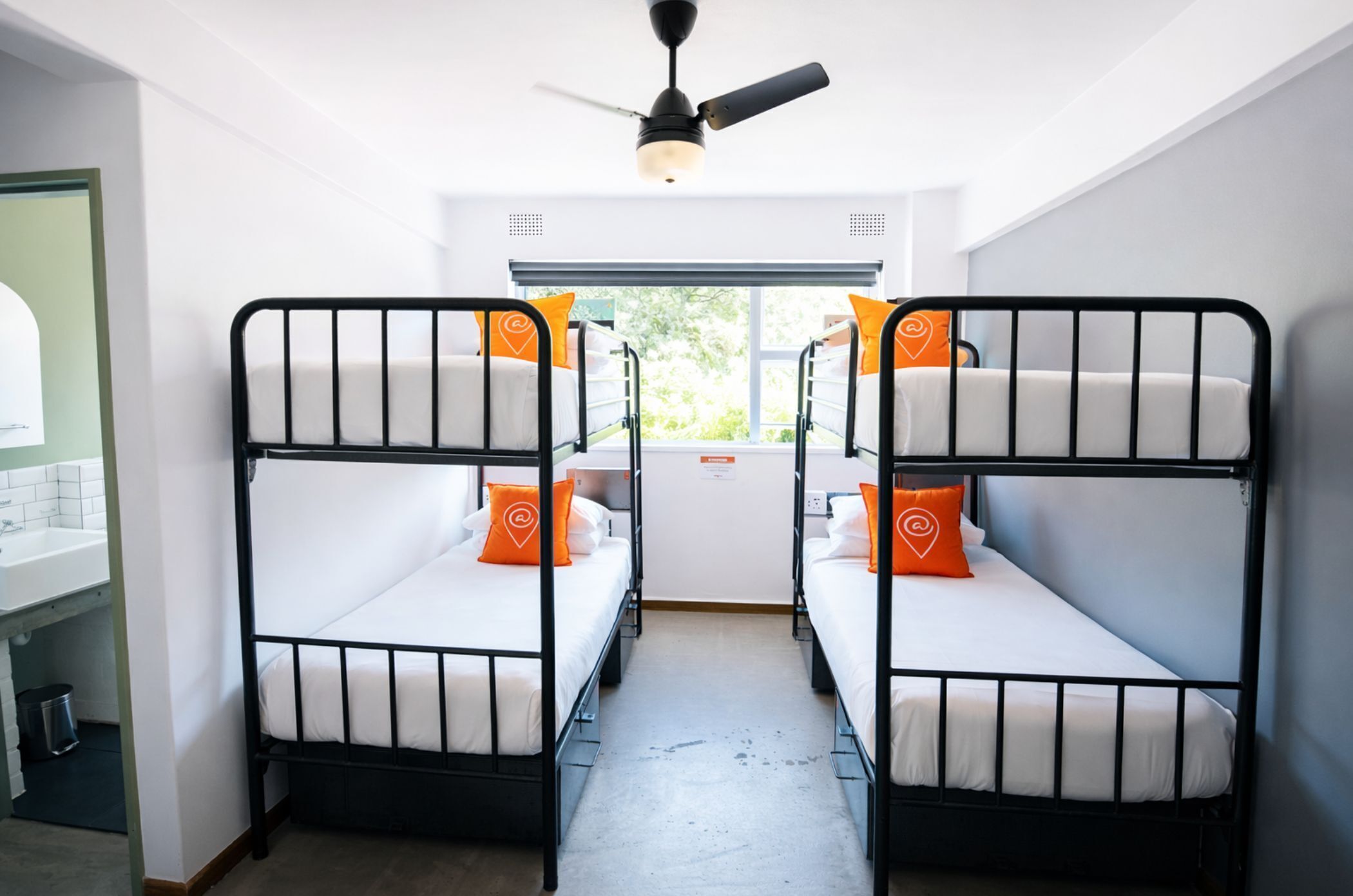 A dormitory room with two bunk beds, a ceiling fan, a window with a view, and a sink in the corner.