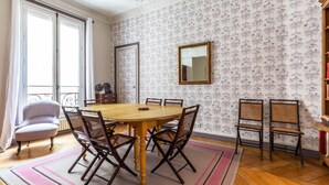 A dining area with a round wooden table surrounded by wicker chairs, a white armchair, and a patterned wallpaper. - Vintage Violet, by Veeve (Paris)