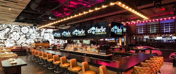 Uma área de bar interior com um balcão comprido, vários bancos e um letreiro de neon com a inscrição 'elotehete'.