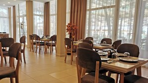 A dining area with multiple tables and chairs, large windows with curtains, and a vase with flowers. - New Akileda Hotel (Golem)