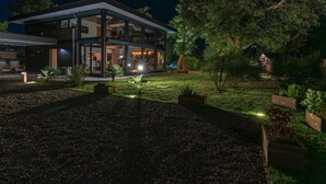 Une maison moderne dotée d’une façade en verre, d’un espace extérieur bien éclairé et d’un jardin où poussent différentes espèces de plantes.
